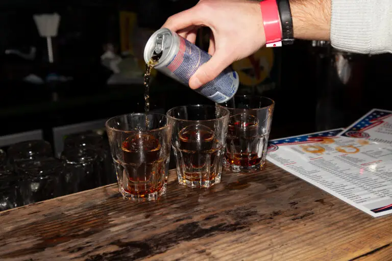The hand of a man wearing a pink wristband pouring Redbull to make three Jagerbombs