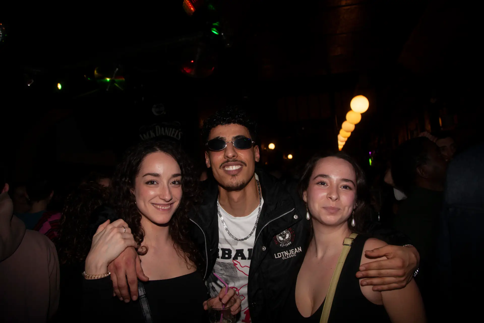 A smiling guy wearing sunglasses holding one girl on each side