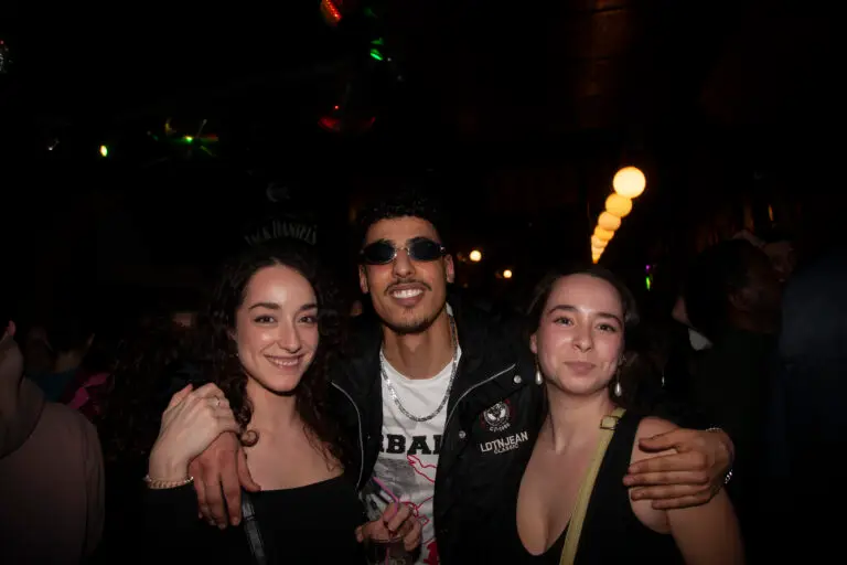 A smiling guy wearing sunglasses holding one girl on each side